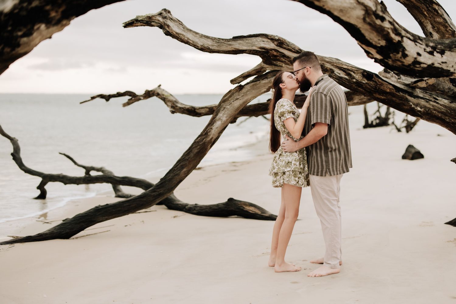 A couple sits on a sandy beach at sunset near driftwood logs along the shoreline.