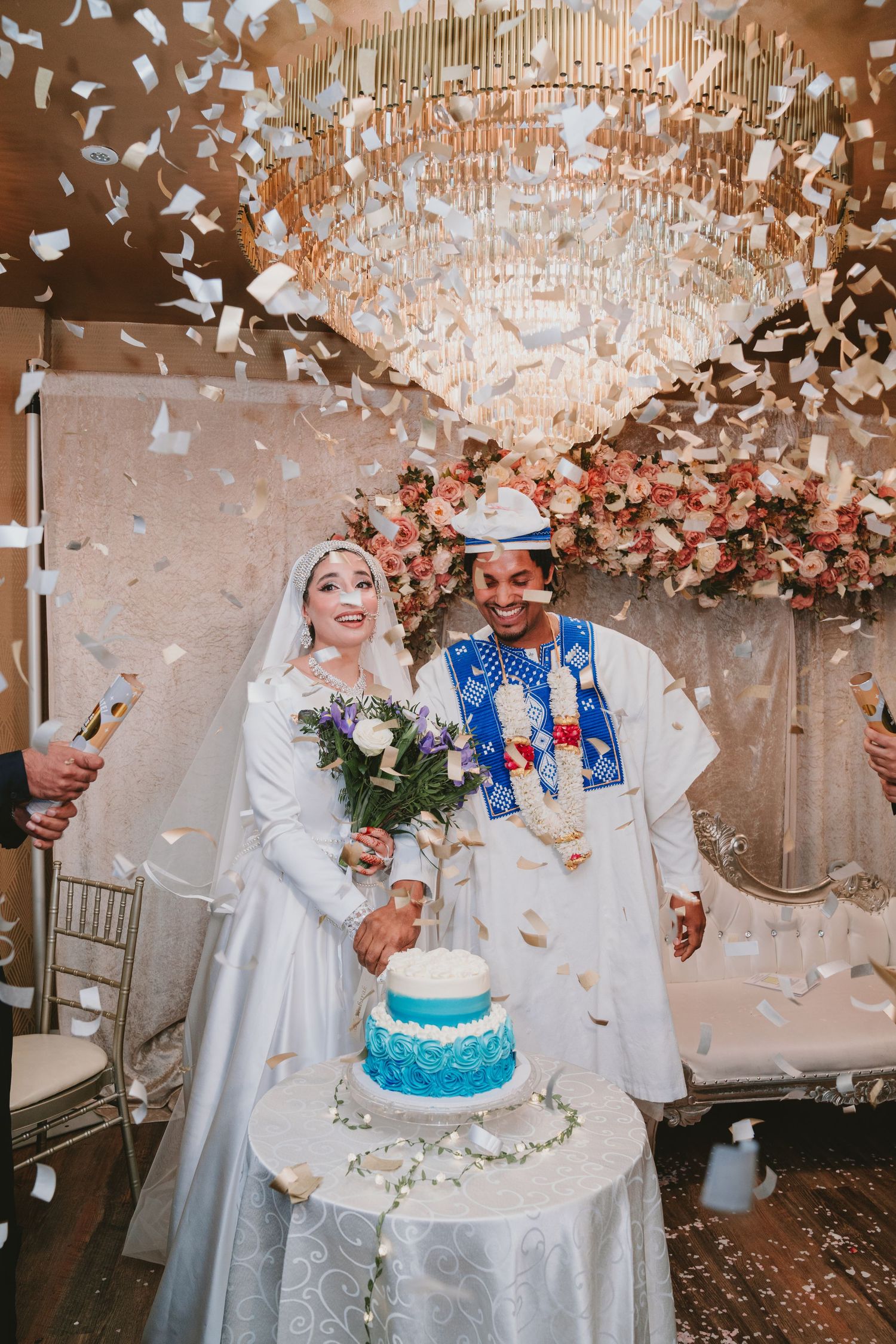 A couple cuts their blue wedding cake under falling confetti and floral decorations.