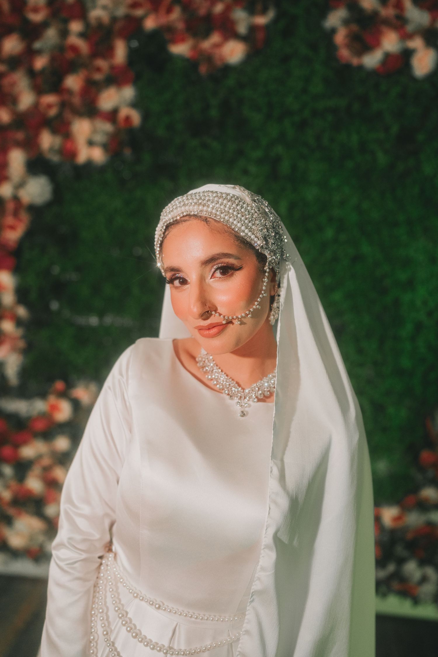 Elegant bridal portrait against floral wall wearing pearl-adorned white dress and veil.
