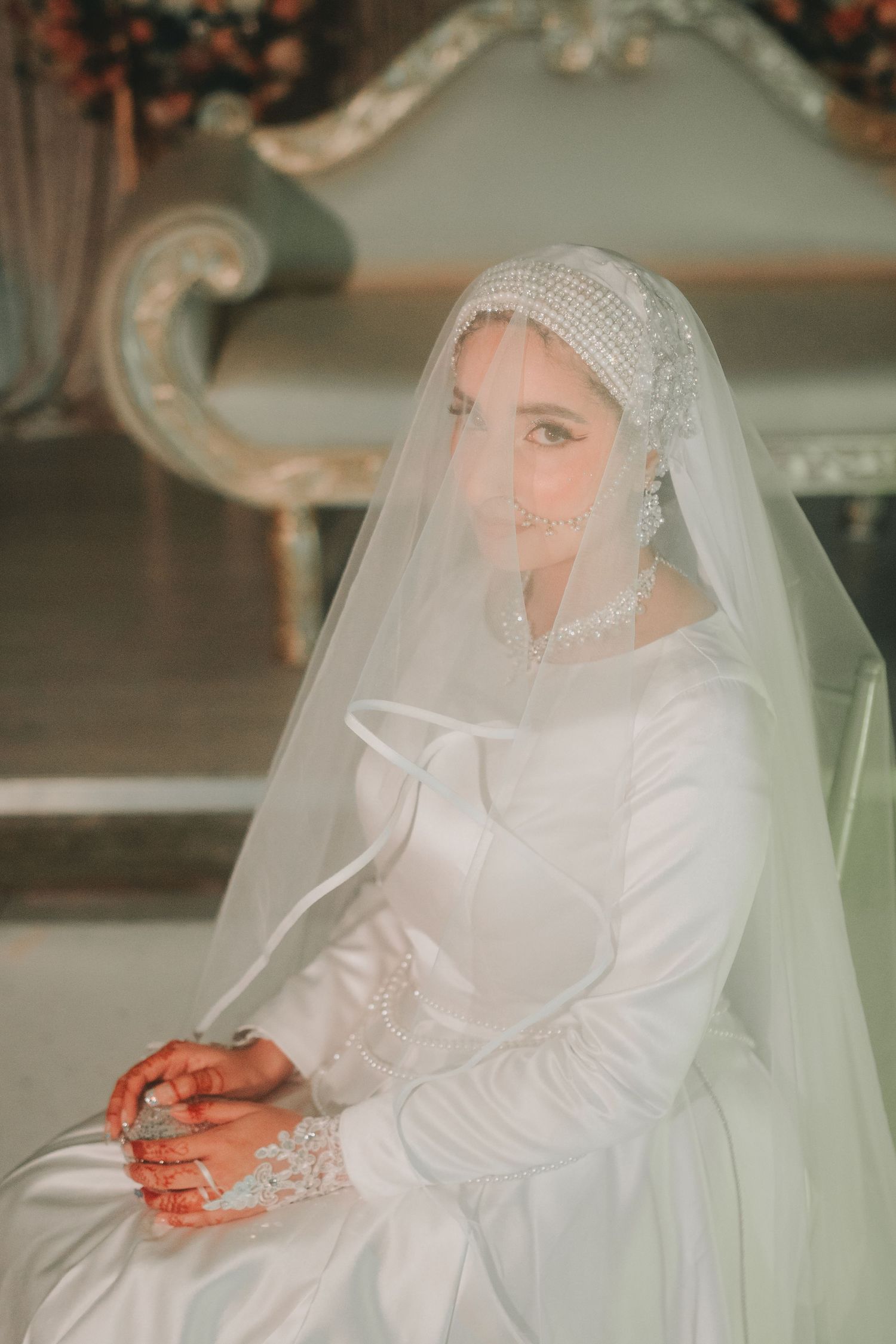 Elegant bride in a long-sleeved white dress and lace veil sits gracefully during her traditional Islamic wedding ceremony.