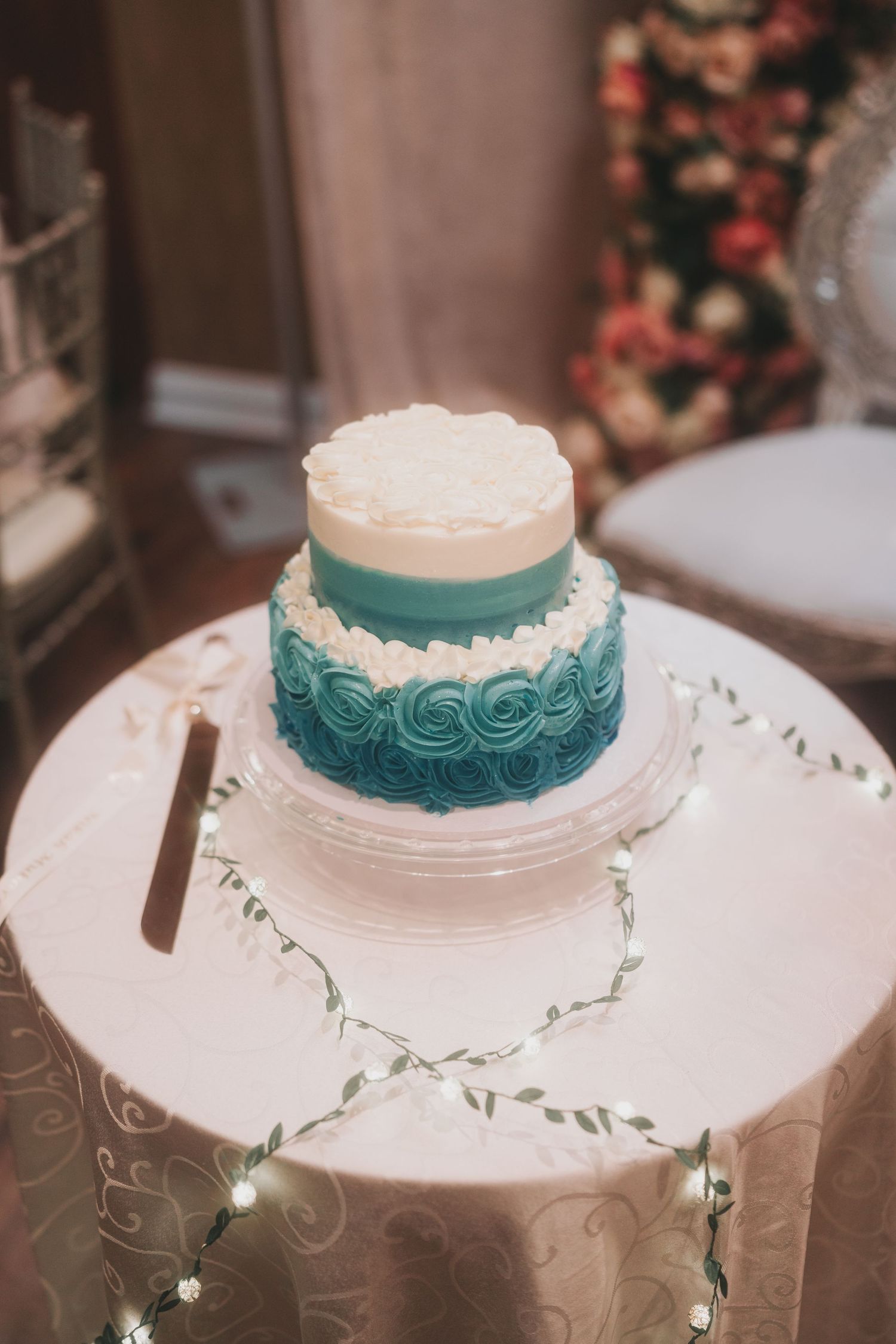 Elegant two-tier wedding cake decorated with turquoise ombre rosettes and pearl details on a white cake stand.