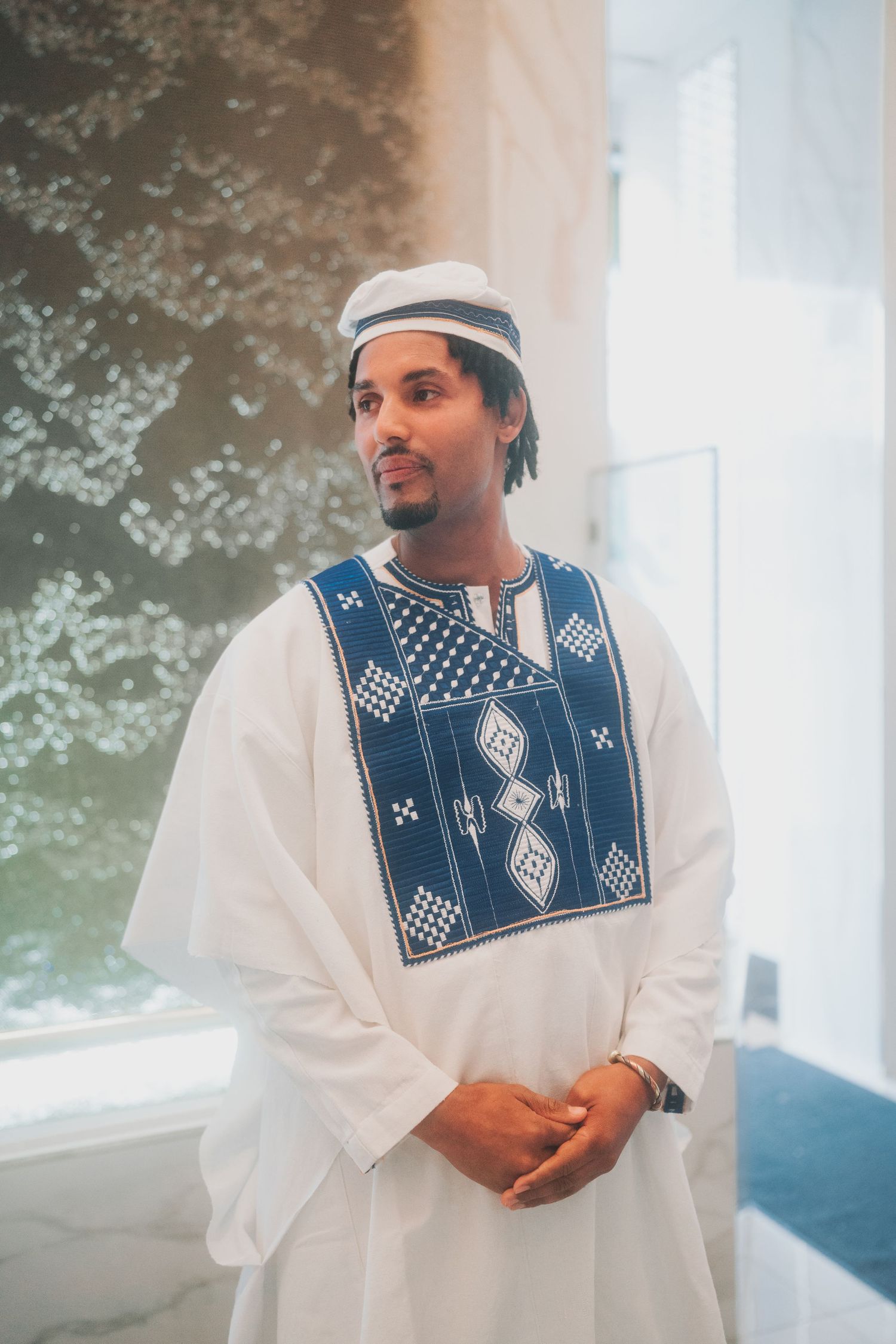 Traditional groom wearing white thobe with blue embroidered vest and head covering at wedding ceremony.