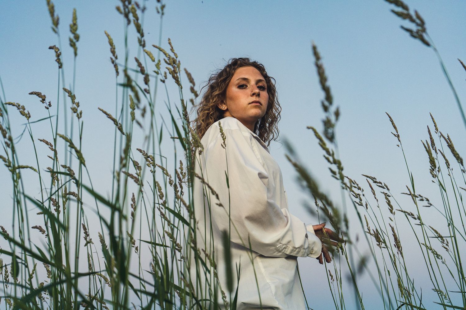 Artistic shot of figure in white clothing among tall grass against blue sky.