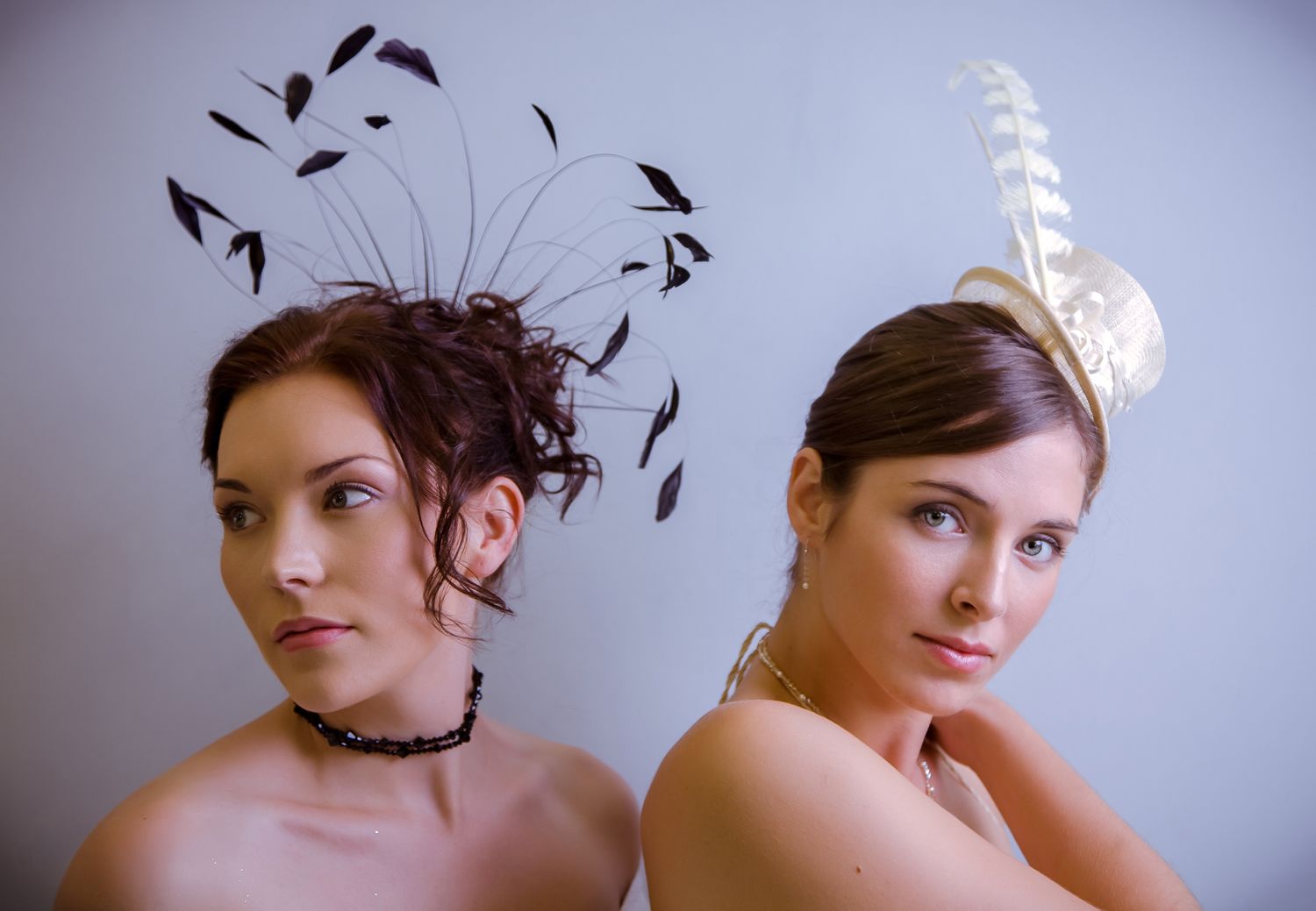 Two people wearing contrasting black feather and white feather hair accessories pose creatively against a plain wall.