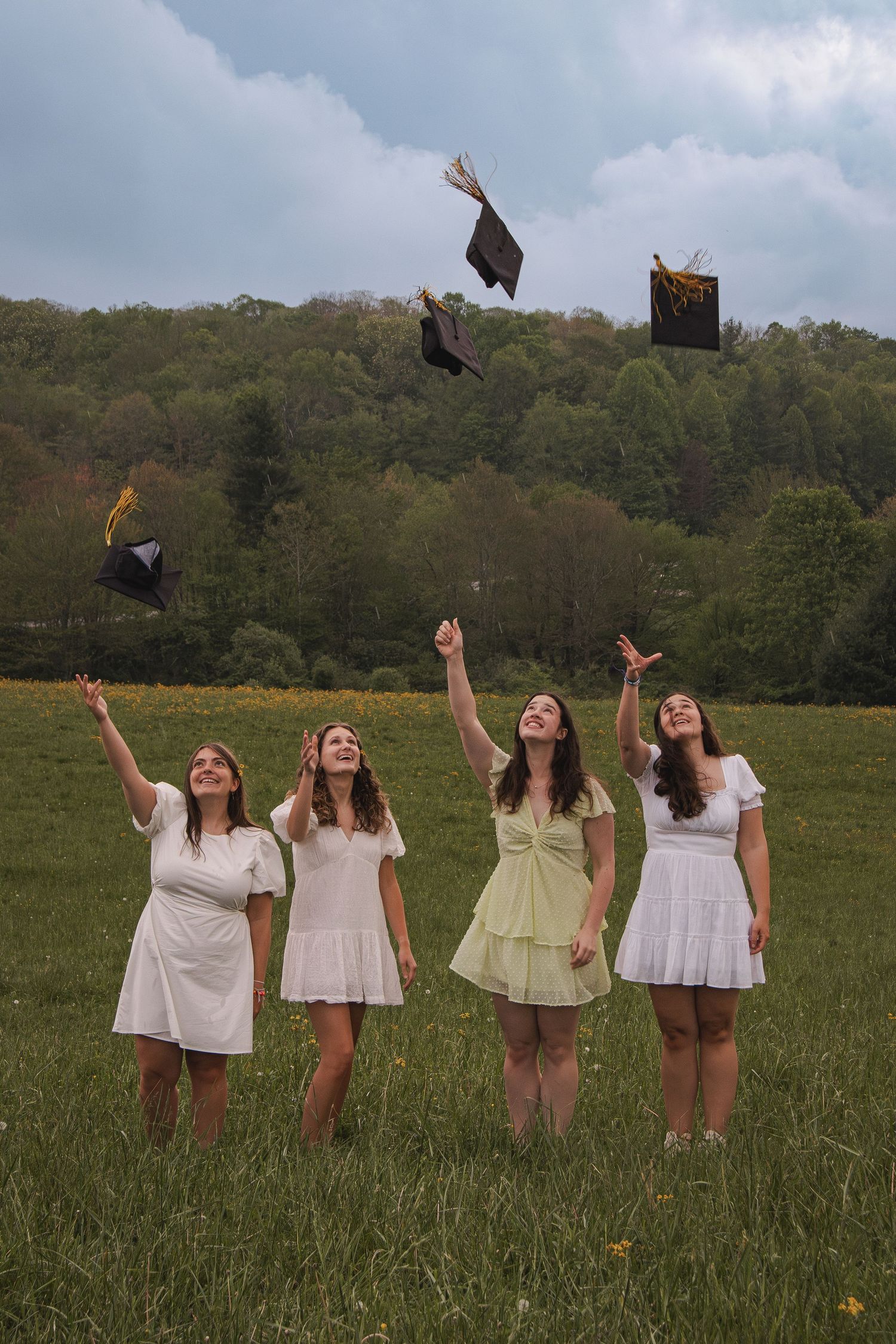 Graduates in white dresses throwing mortarboards into the air in a sunny field. Luxury wedding photographer North Carolina