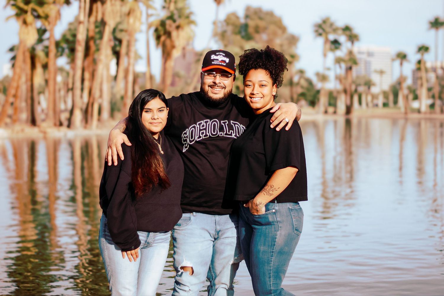 Three friends pose together by palm tree-lined lake during sunset.