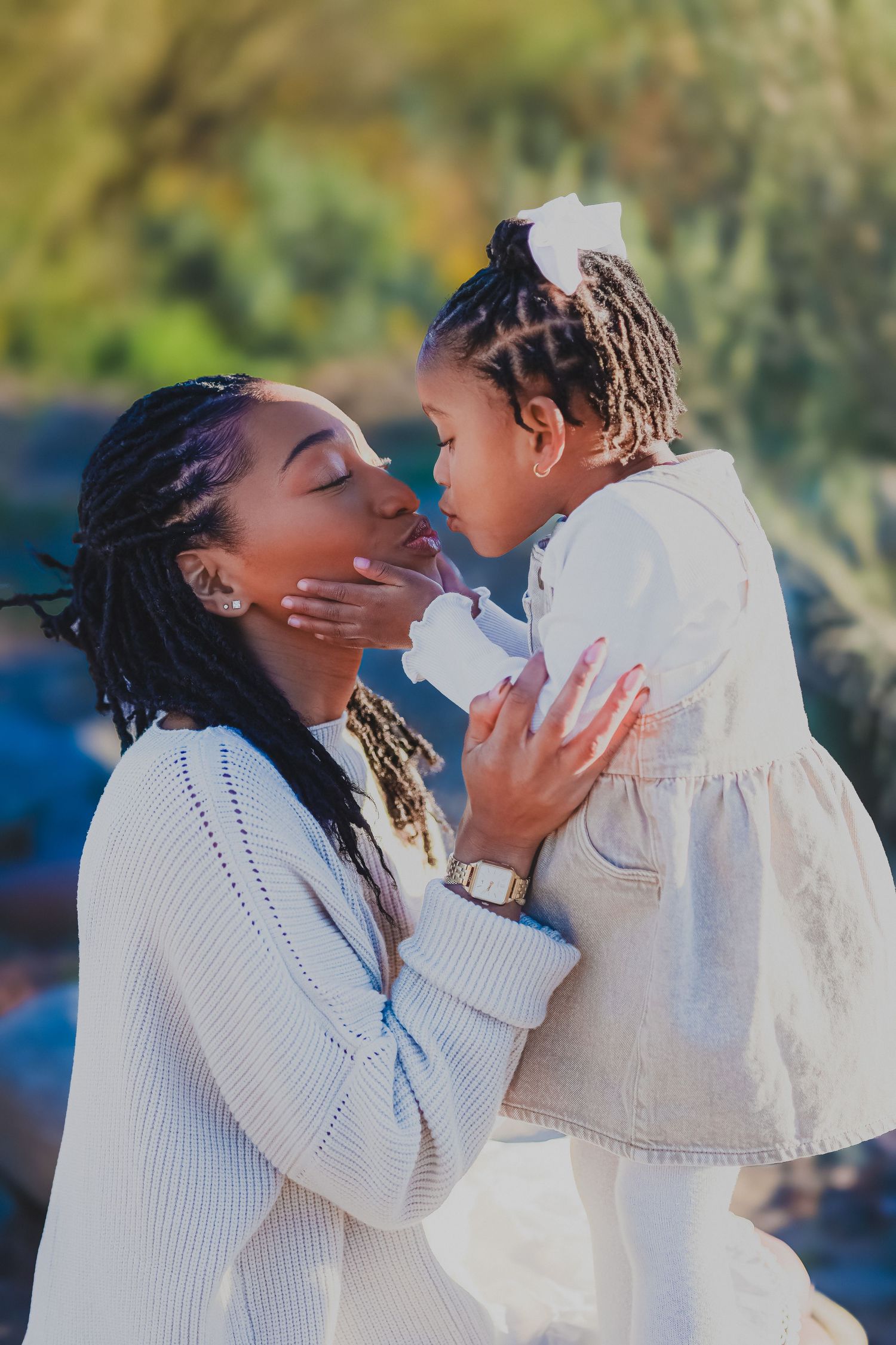 A mother with braids holds her toddler daughter close as they share a tender nose-to-nose moment outdoors.