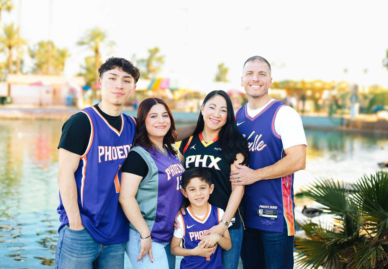 A family wearing Phoenix Suns basketball jerseys poses together by a waterfront during golden hour.