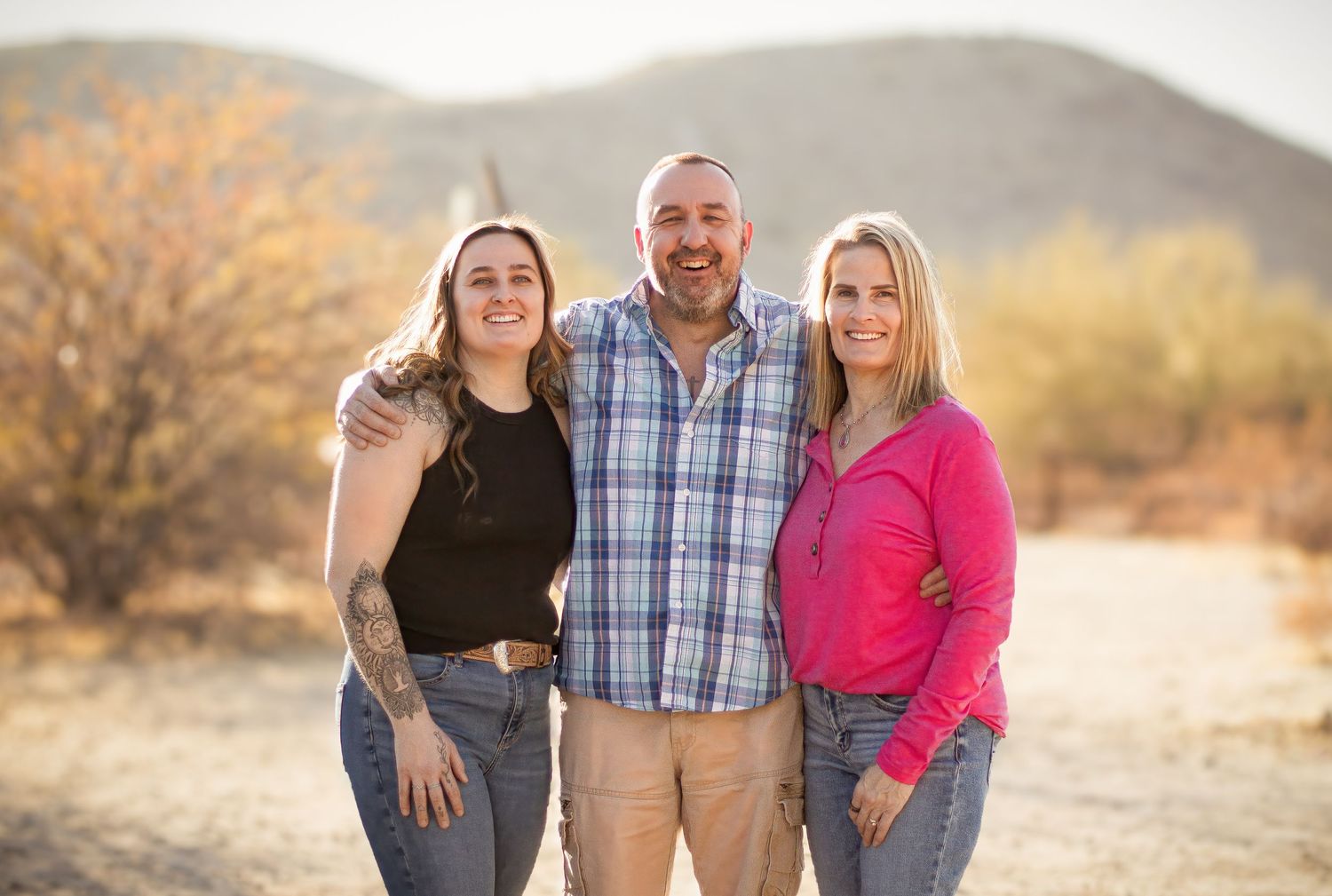 Three people pose together outdoors with desert mountains in the background at sunset.