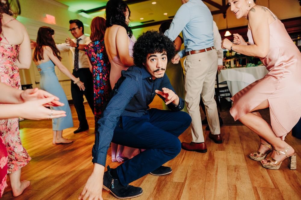 Party guests dance on a polished wooden dance floor while someone in a blue outfit crouches low with a camera.