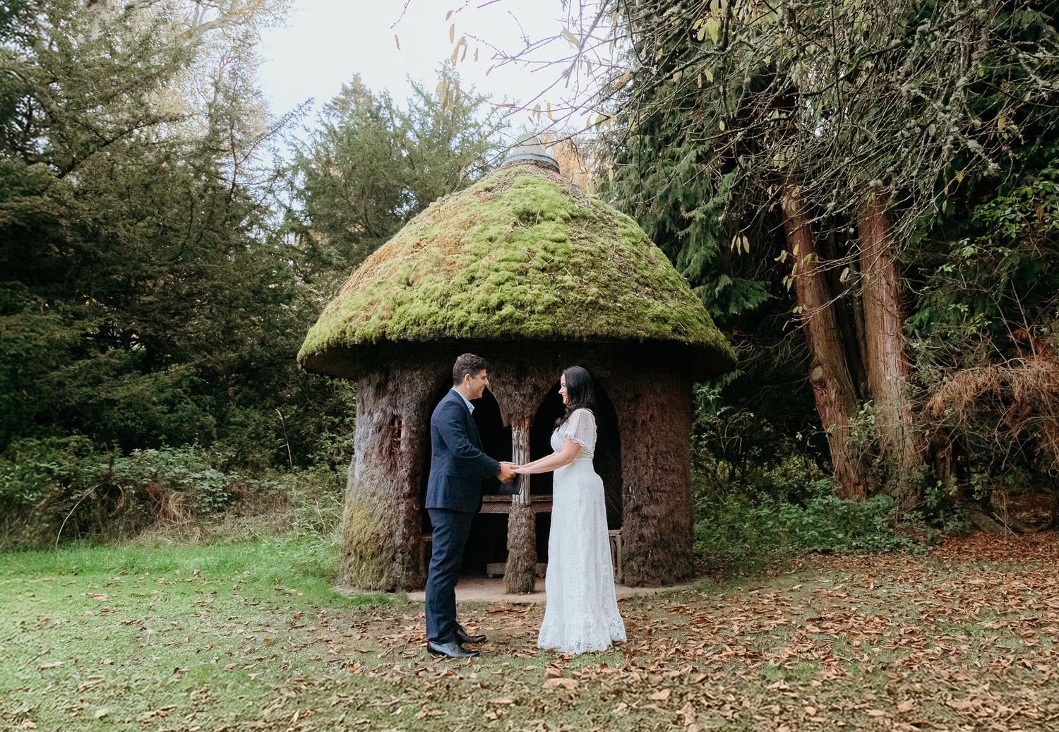 A moss-covered thatched gazebo stands in a wooded area surrounded by autumn leaves.