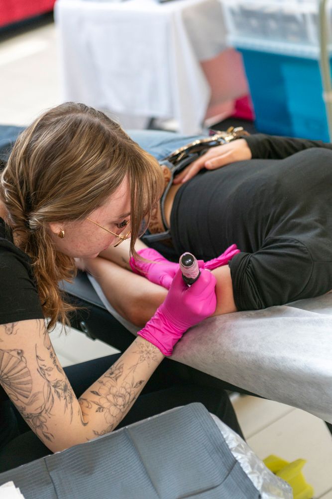Female tattoo artist with pink gloves and tattooed arms carefully inking a client's arm during a tattoo session.