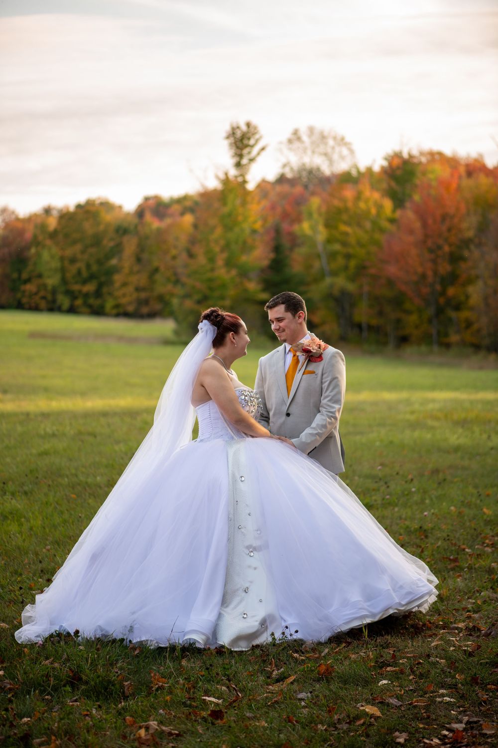 Couple in wedding attire sitting together on autumn grass with colorful fall foliage in background.