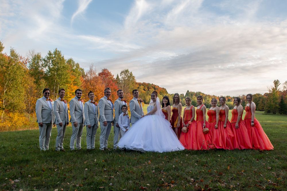 Wedding party in formal attire poses in a scenic outdoor setting with bridesmaids in red dresses and groomsmen in gray suits.