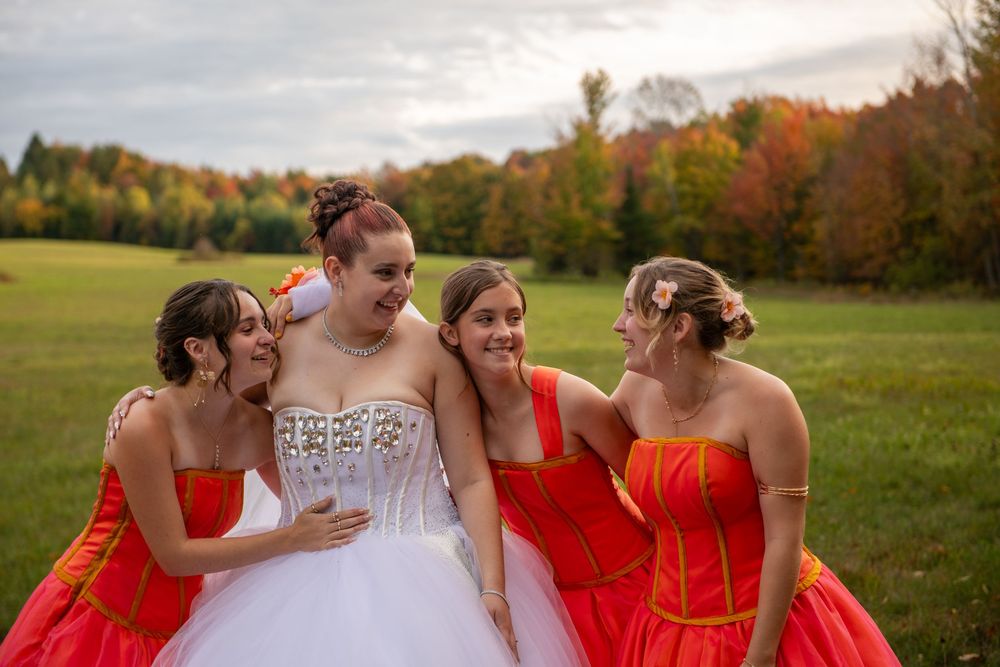 A wedding party in a scenic autumn field with bridesmaids wearing bright red dresses and a bride in white.