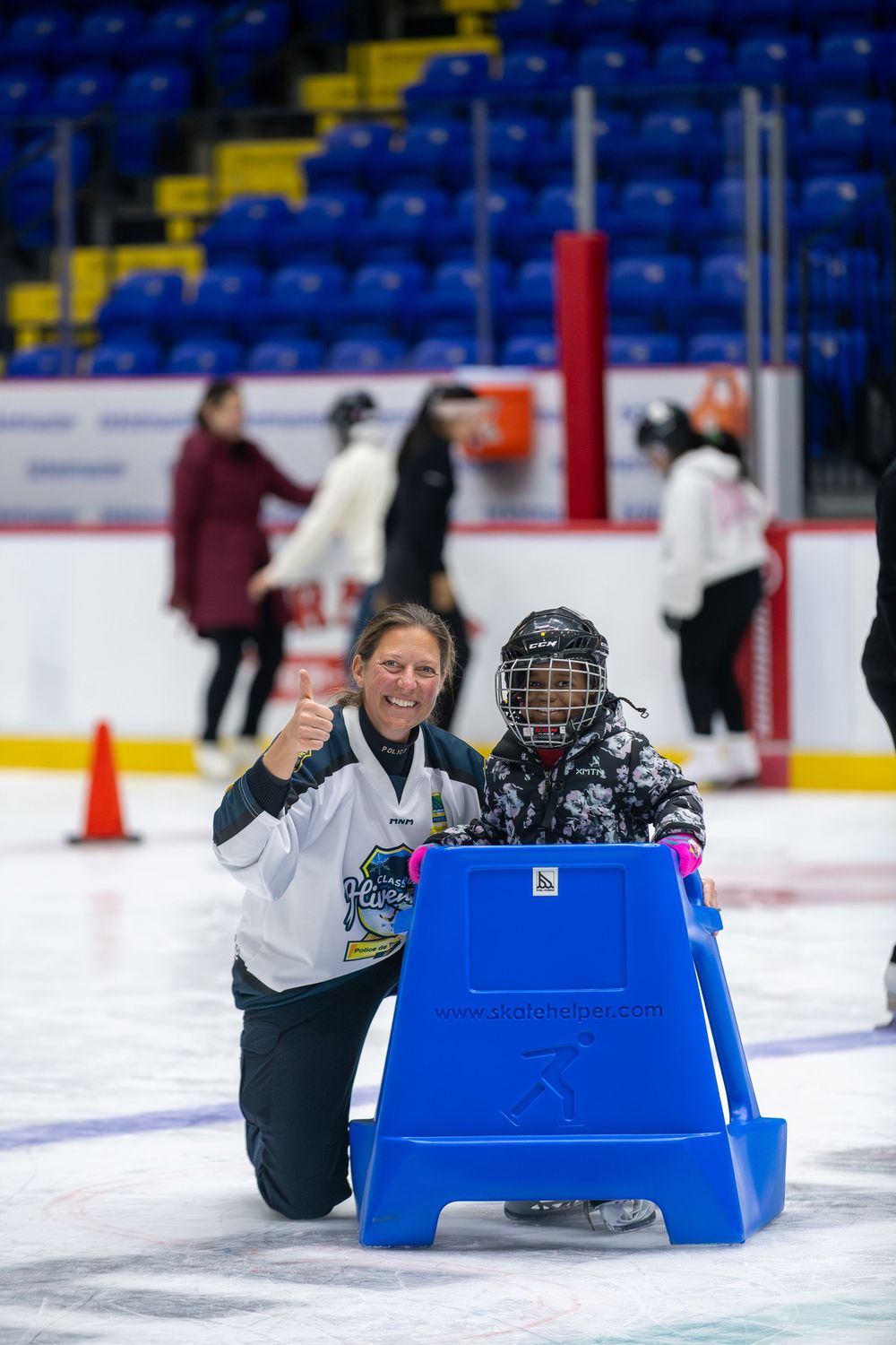 A young skater uses a blue training aid while learning to ice skate at an indoor rink.
