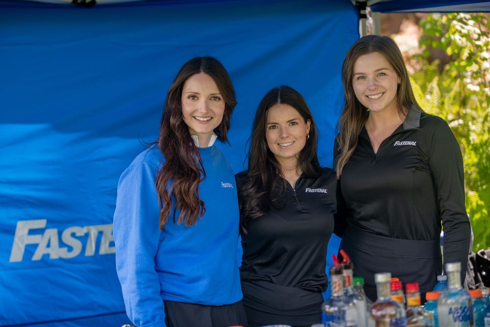Three women smiling together at a Fastenal sponsored outdoor event, wearing branded polo shirts and jackets.