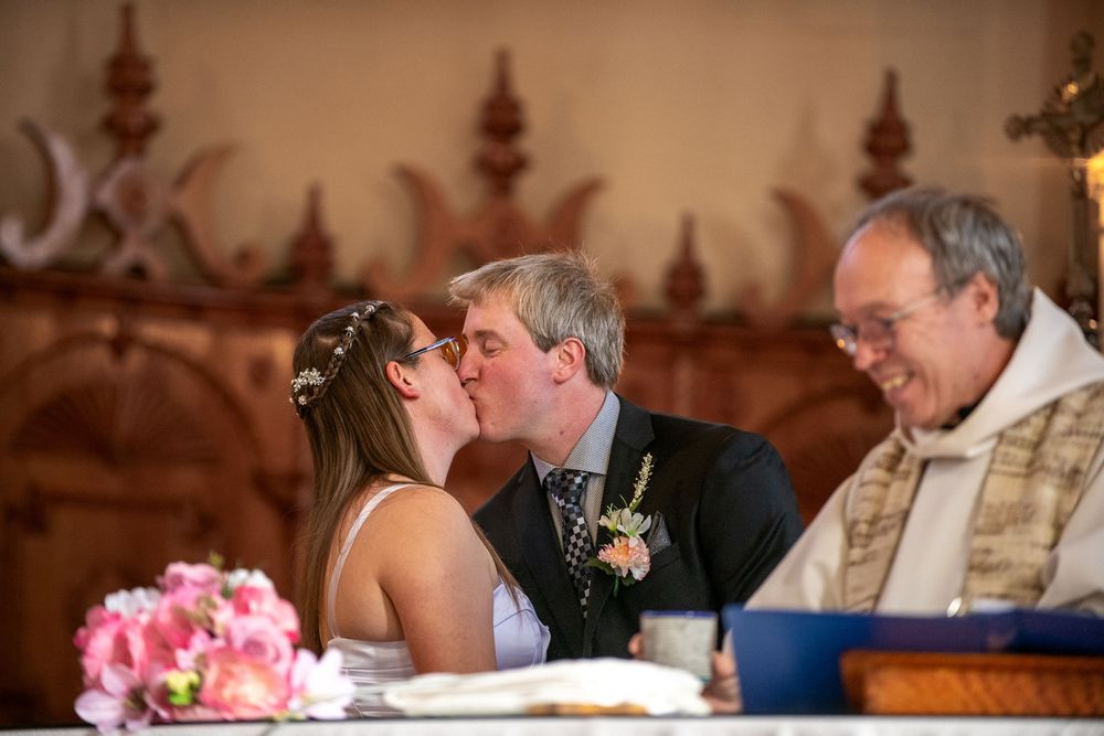 A couple shares their first kiss at the altar during a church wedding ceremony with pink floral bouquet in view.