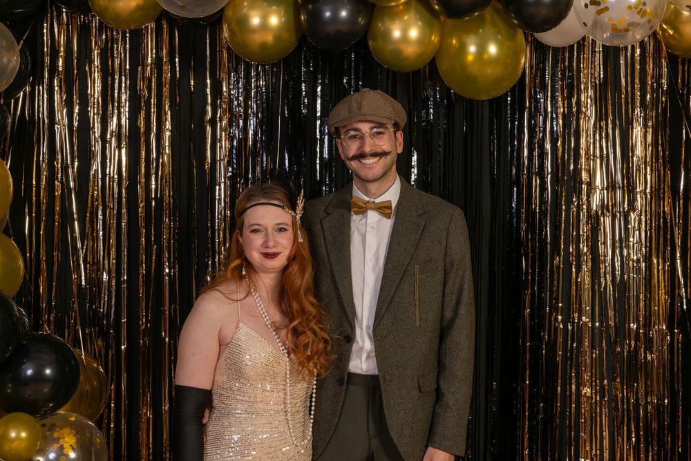 Couple poses in front of gold glittery curtain backdrop at formal event with festive gold balloons overhead.