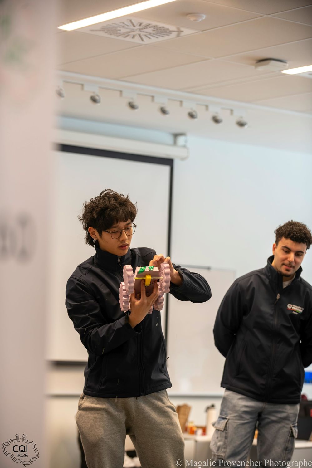 A person in a black jacket demonstrates a sequence of hand movements while holding small objects in a classroom setting.