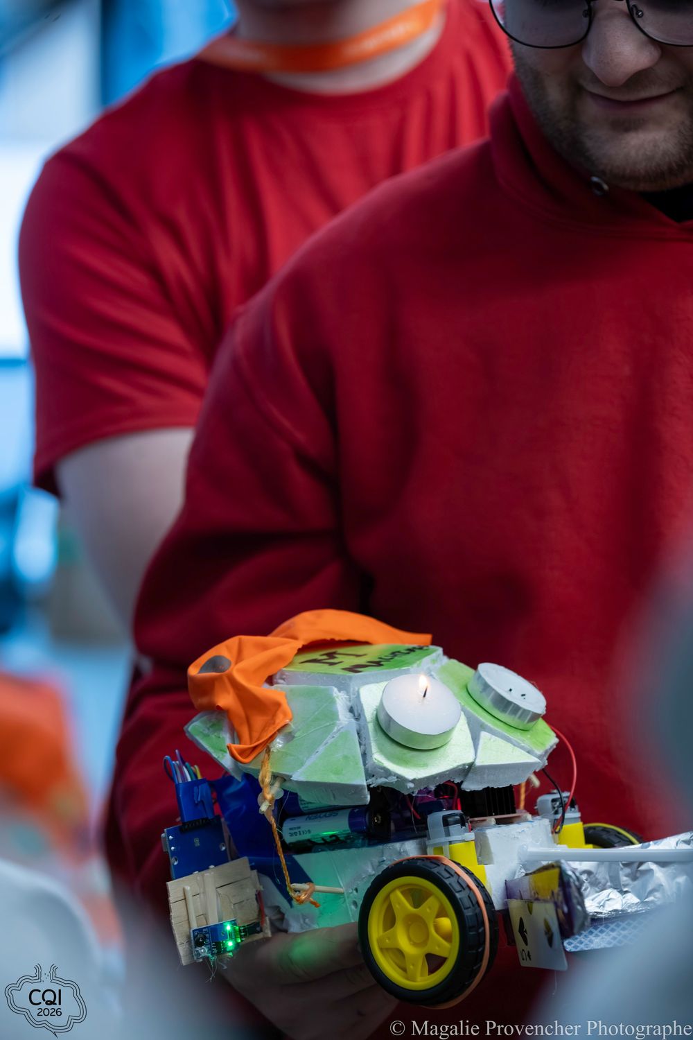 A person in a red shirt holds a colorful robotic toy made of plastic building blocks with yellow wheels.
