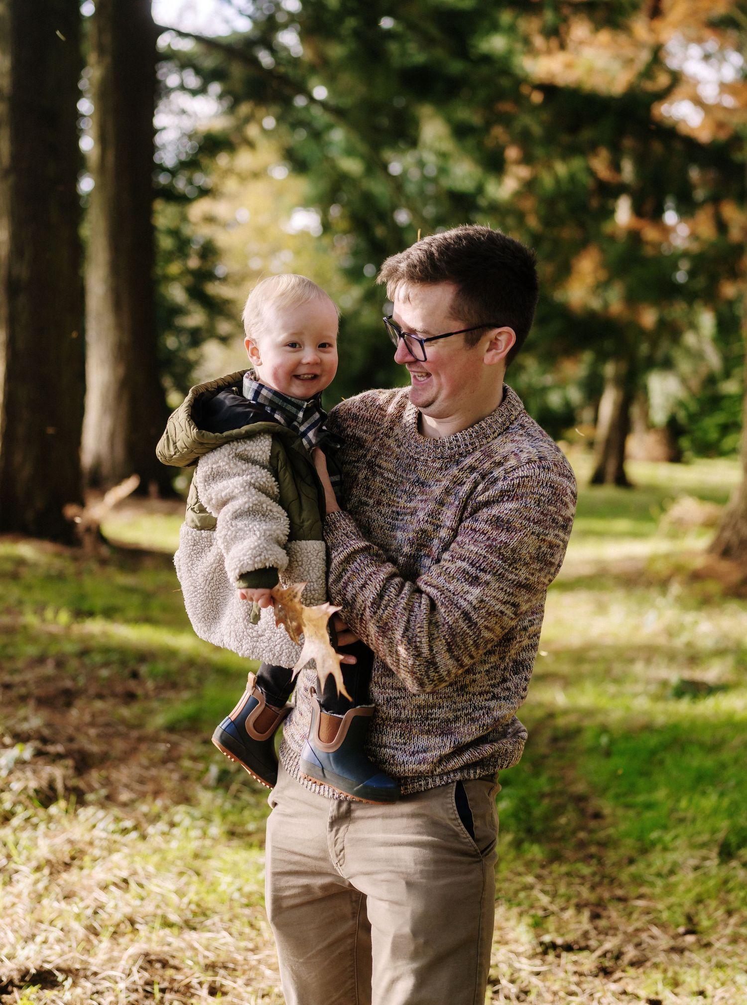 A father holds and laughs with a baby while standing in a wooded outdoor setting on a sunny autumn day.