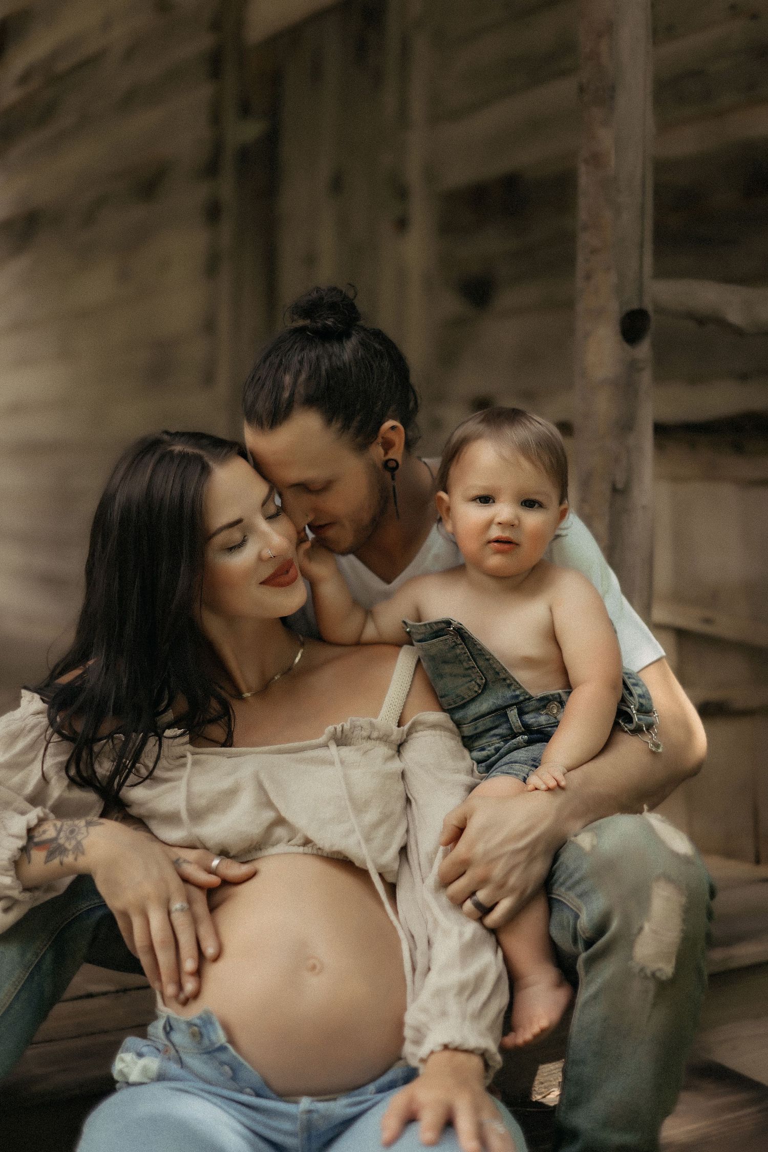 Intimate maternity photo shows expecting parents and toddler in rustic setting sharing tender family moment.