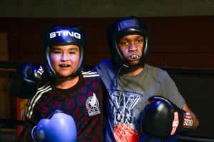Two young boxers wearing protective headgear stand together in a gym setting.