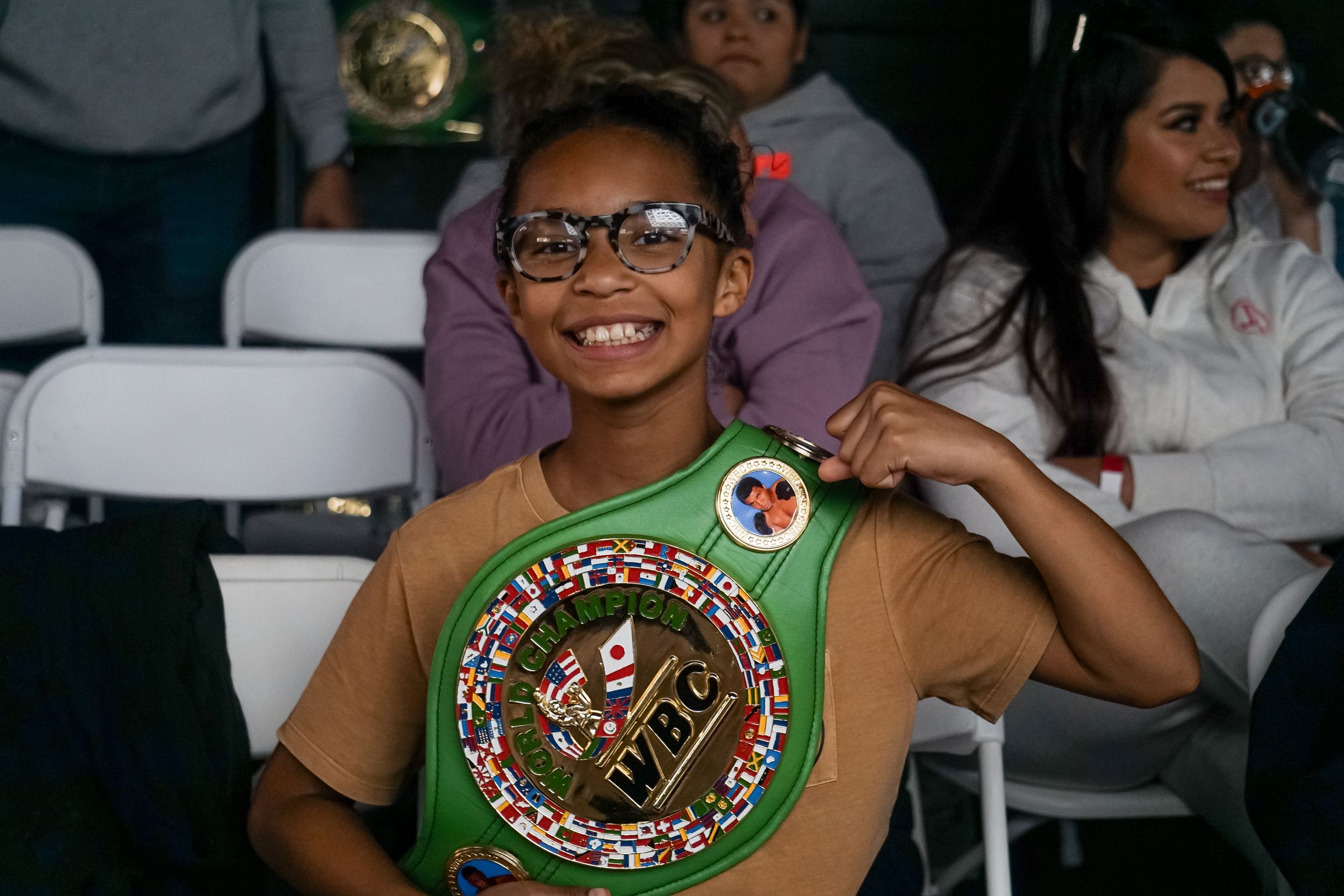 A young person wearing glasses and a green sash with a decorative medallion sits smiling in an audience.