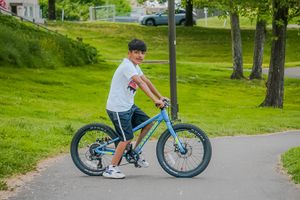 A cyclist in a white t-shirt rides a turquoise mountain bike on a paved path through a green park setting.