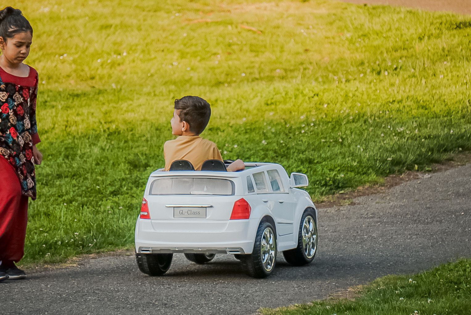 A child rides in a small silver toy luxury car on a path while someone walks alongside on a grassy lawn.