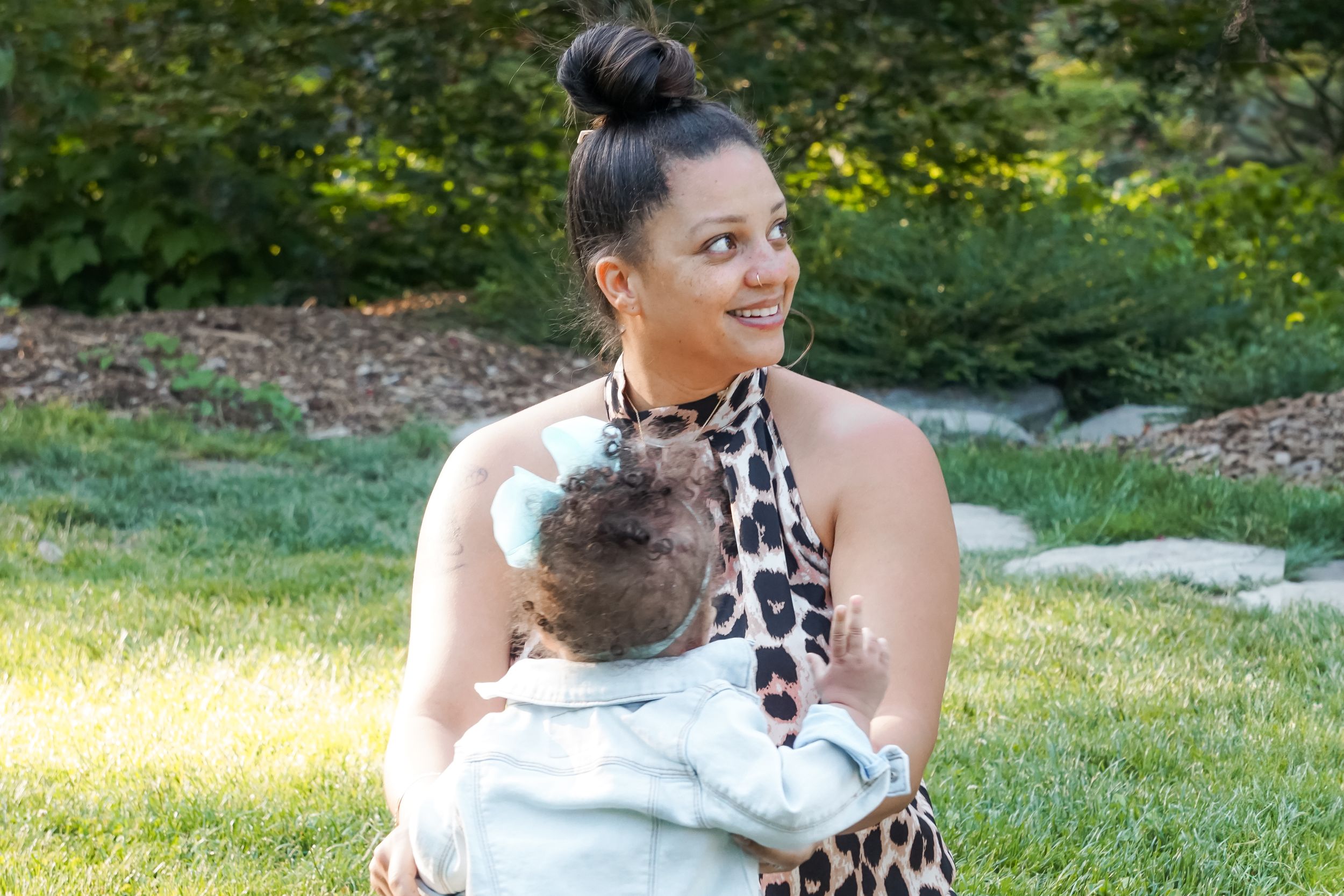 A person wearing a leopard print dress holds a baby while sitting in a sunny garden area.