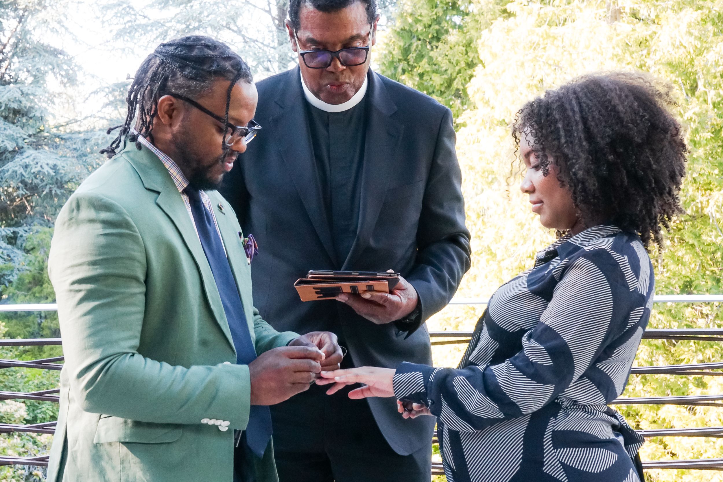 A couple exchanges rings during an outdoor wedding ceremony with an officiant reading from a tablet.