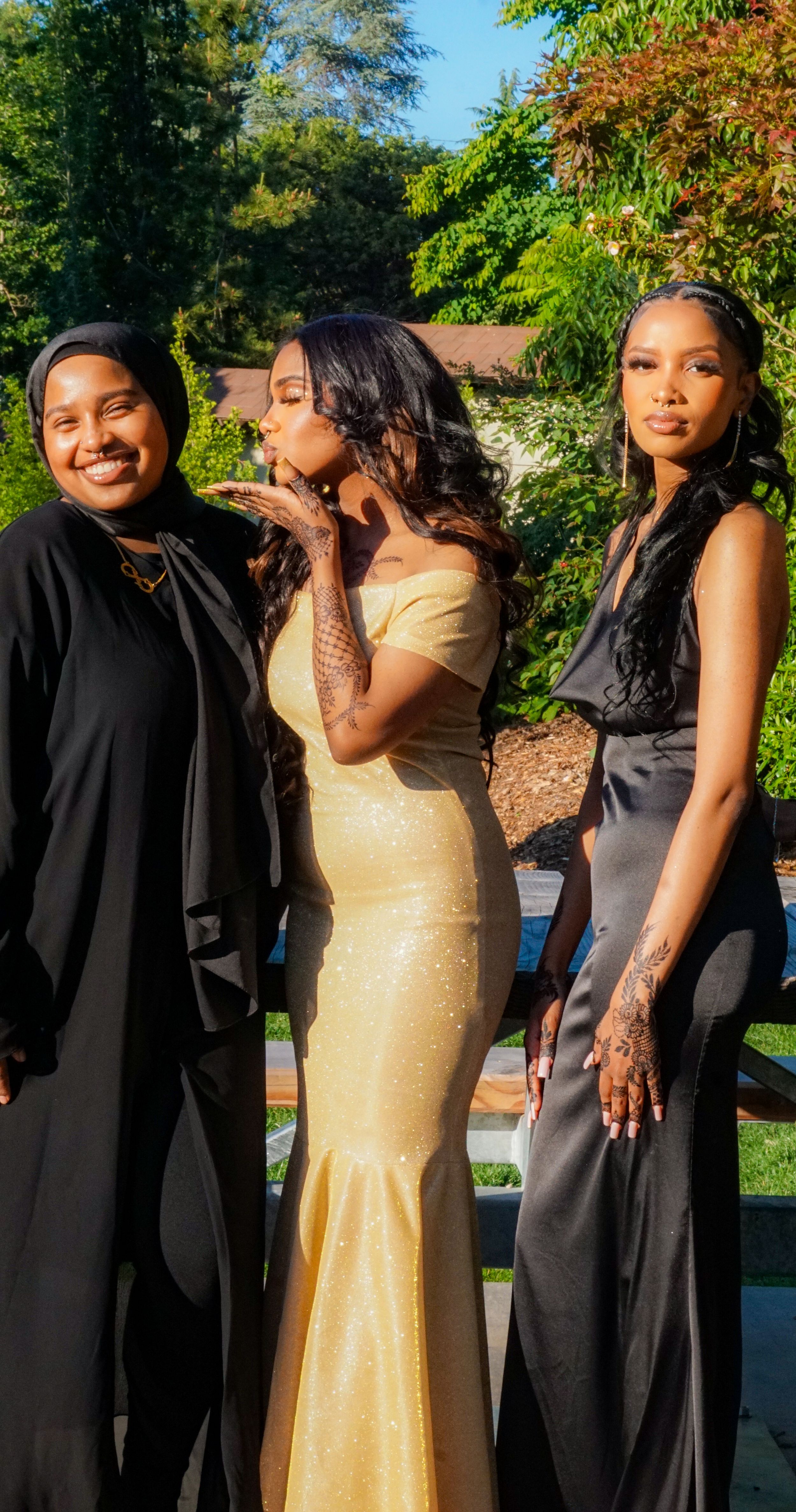 Three people in formal attire pose together outdoors, wearing a combination of black and gold dresses.
