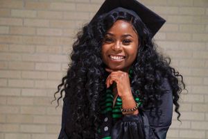A graduate in cap and gown with curly hair poses against a brick wall background.