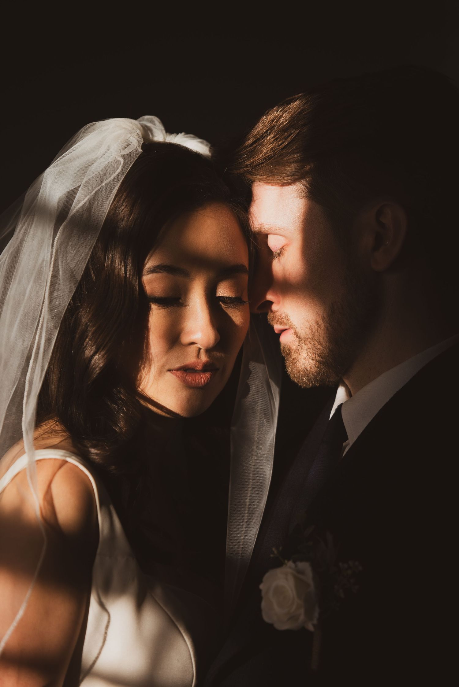 Intimate wedding portrait of couple in dark moody lighting with bride in white veil and groom in black suit.