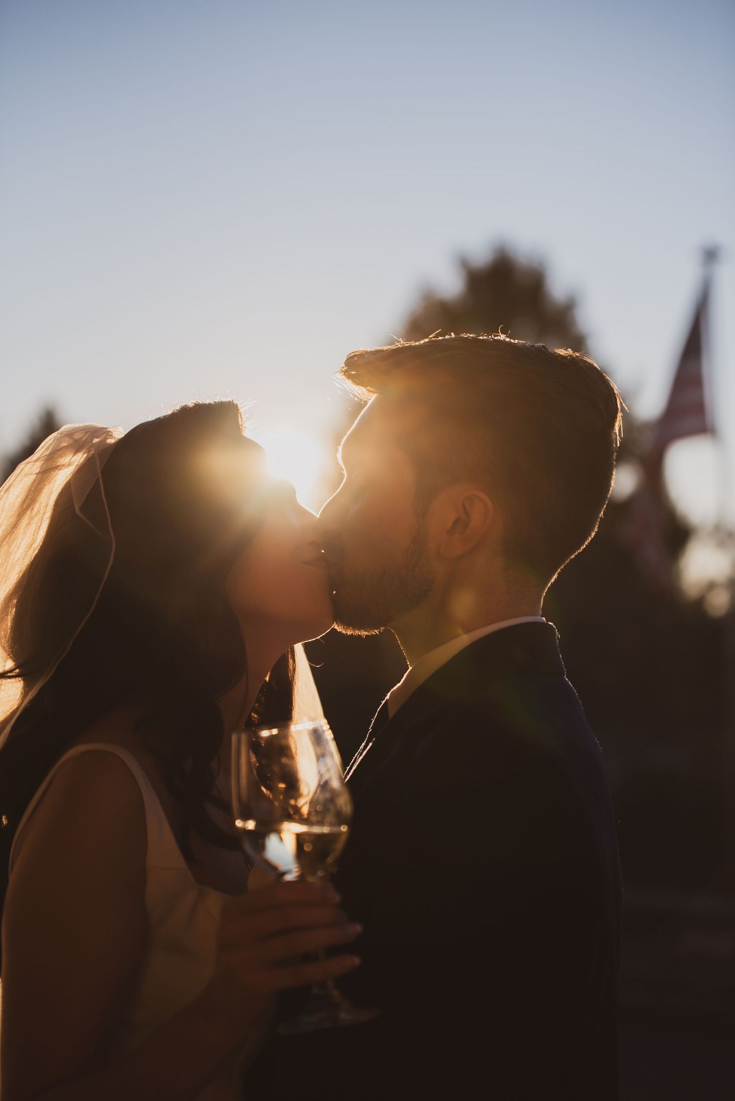 Silhouetted couple sharing romantic sunset kiss while holding champagne glass with church spire in background.