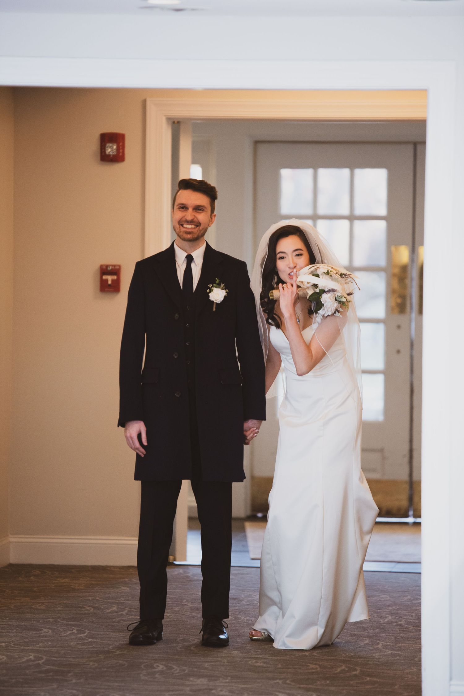 A couple in formal wedding attire stands in a hallway with white doors and beige walls.