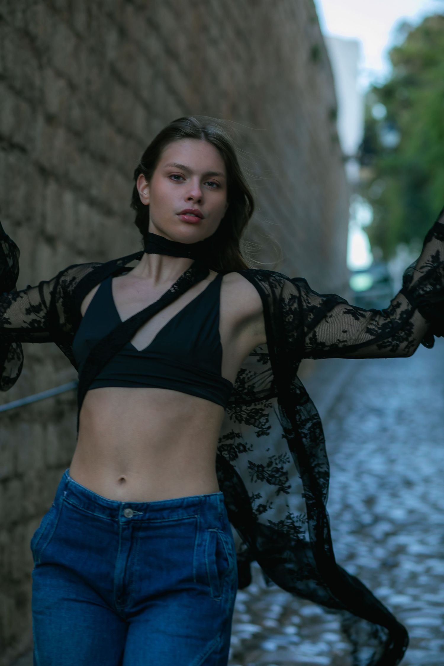 Model wearing black lace top and jeans poses against stone wall in moody outdoor setting.