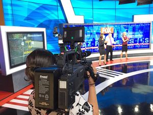 Camera operator films a news broadcast in a television studio with blue backdrop and display screens.