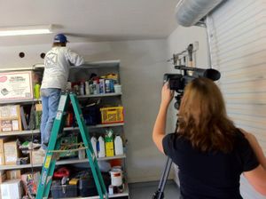 Camera operator films while standing on a ladder in a storage room with shelves.