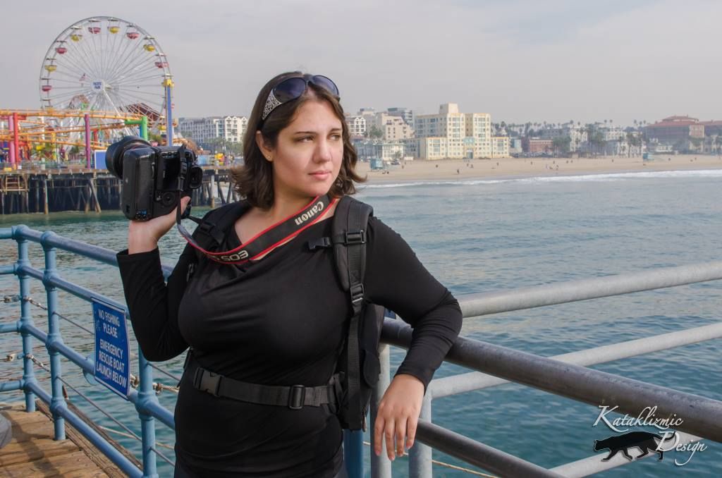 Videographer with camera equipment stands on pier with ferris wheel and beach in background.