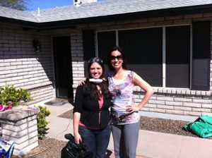 Two women pose together outside a white brick house on a sunny day.