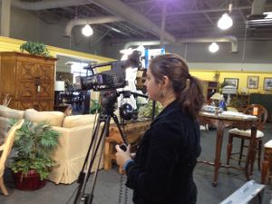 A camera operator films in a furnished room with lighting equipment visible.