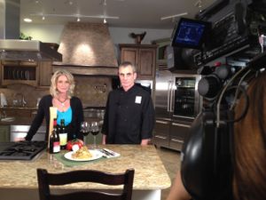 Camera filming a cooking segment with granite countertops and upscale kitchen cabinetry in background.