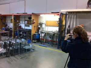 Camera operator filming in industrial workspace with equipment and fluorescent lighting.