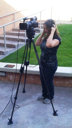 Camera operator adjusts equipment while filming outdoors on a tripod.