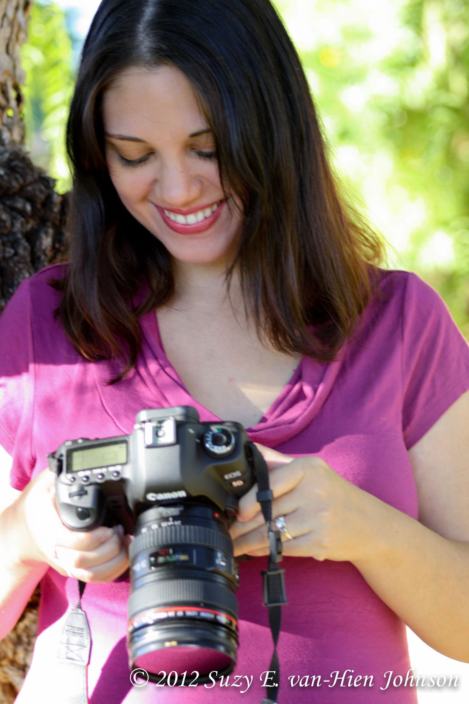 Photographer in pink shirt smiles while reviewing images on a DSLR camera outdoors.