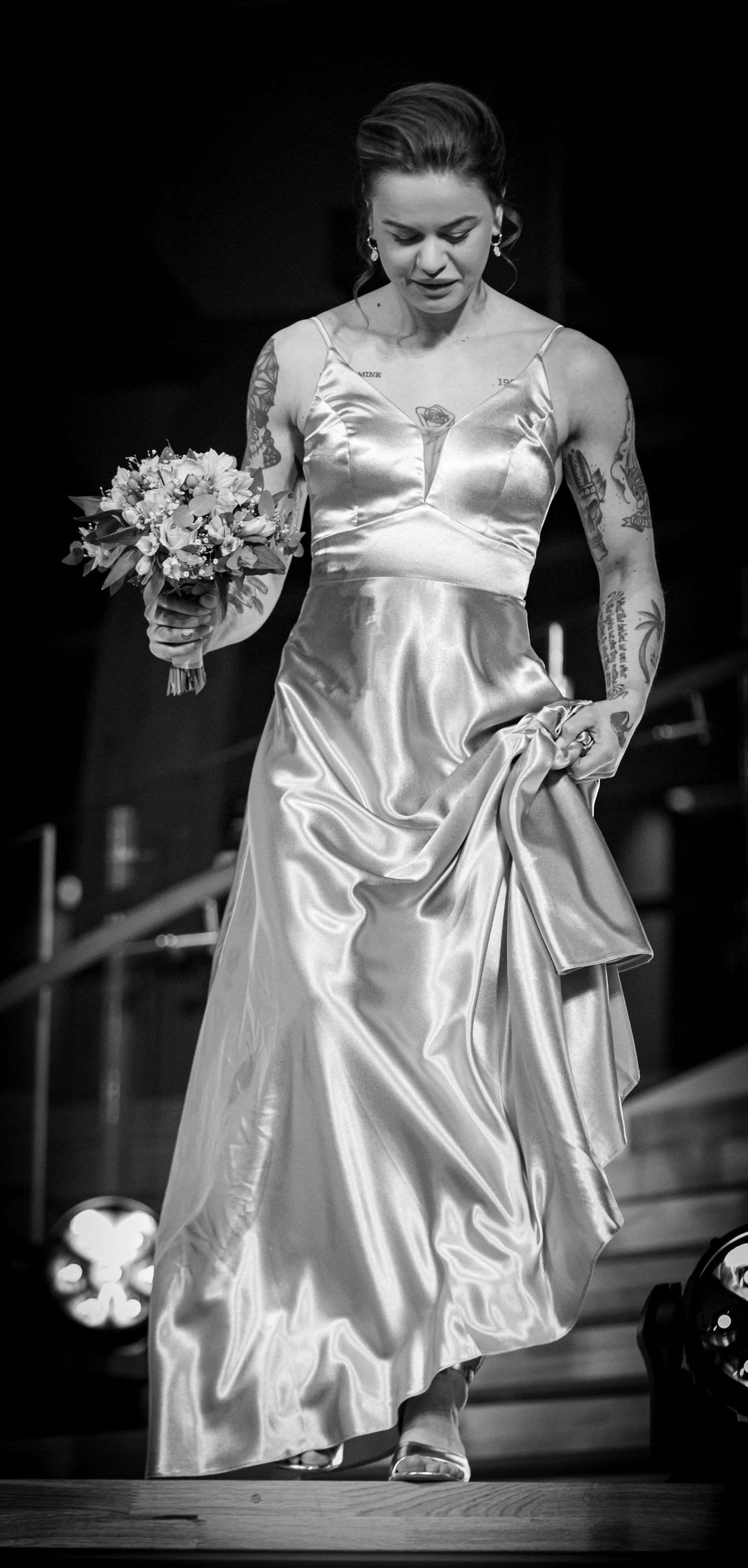 Black and white full-length photo of tattooed bride in satin gown holding a bouquet.
