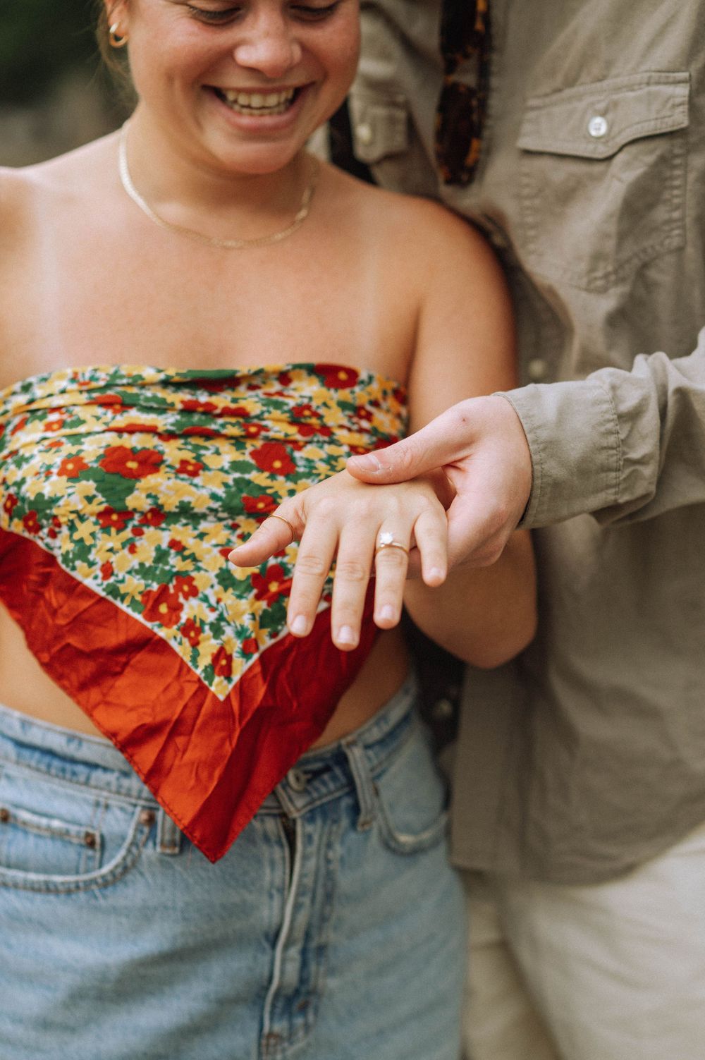 A newly engaged couple hugging after a secret proposal, facing the camera and showing their hands. The woman’s engagement ring is visible.